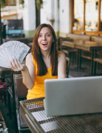 woman-outdoors-street-coffee-shop-cafe-sitting-with-modern-laptop-pc-computer-holds-hand-bunch-dollars-banknotes